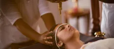 Woman lies on her back with her eyes closed as oil drips onto her scalp and a therapist holds her head