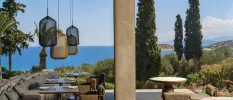 Open-sided restaurant terrace with cream sofa seating, hanging pendant lights and a view of the ocean and surrounding green hillsides