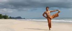 A topless Thai man in loose orange trousers on tiptoe holding his other outstretched leg, on Nadan Beach under a cloudy sky