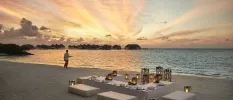 Low white L-shaped table laid up with a white tablecloth and candles, surrounded by cushioned benches on a sandy beach at sunset
