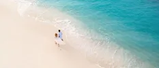 Couple in white strolling hand in hand on a white-sand beach, with vivid turquoise waves lapping against the shore