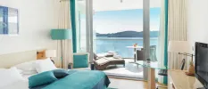 Modern hotel bedroom with white walls and bedding, blue soft furnishings, a desk with a TV and a furnished balcony with a sea view