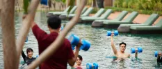 Staff member in red leads a class of people in a swimming pool holding up blue dumbbells