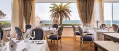 Bright restaurant with wooden floors, cream drapes and large windows with an ocean view, plus tables with crisp white tablecloths and elegant dark chairs