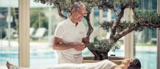 Smiling woman lies on a bed as a staff member in a white polo shirt smiles down at her, in front of a swimming pool and olive tree
