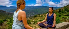 Two women in active wear in lotus position with their eyes closed and green hills in the background