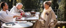 Group in loose fitting clothes sit around a table in the sunshine on a terrace with views of gardens