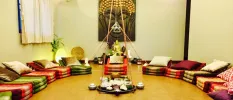 Room with brightly coloured cushions laid out in a circle, a large Buddha picture on the wall and a central set-up for some sort of ceremony