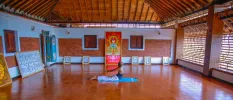 Man practices yoga on a blue towel in a studio with terracotta tiled floors, wood-beamed ceilings and colourful Hindu artworks