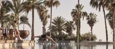 Man dives into a swimming pool edged by palm trees, as a woman in a lime swimsuit watches on