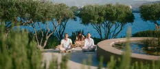 Three people in loose fitting clothes sit with their eyes closed and hands on their knees in a pretty garden with trees, a stone-walled pond and the ocean in the background