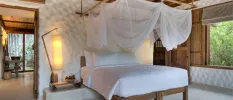 Villa bedroom with a wooden bed, wall panels and ceiling, a stone white wall, white bedding and sheer white drapes hung over the bed, plus lantern-style lighting 