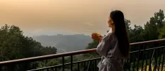 Woman in a white robe holding a yellow mug, overlooking the misty mountains as the sun rises in the distance 