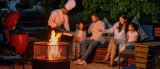 Man, woman and two daughters smile as they sit around a fire at night and a chef serves them from a barbecue