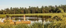 Man in camo attire fishes on a wooden jetty in a lake surrounded by lawns and woodland