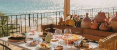 Table laid with rustic wooden bowls, glasses of rose, and a shelf full of pots next to a wrought-iron balcony with a sea view