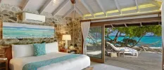 Stylish room with a stone wall, white bedding, blue cushion and throw and seaside painting overlooking and ocean-facing wooden patio with loungers