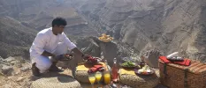 A staff member laying out a mountaintop picnic, with glasses of orange juice, a bread basket, and plates of cheese and salad