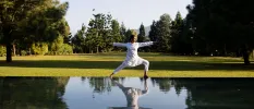 Woman in white loose clothing in a yoga pose in a green field, next to water mirroring her reflection