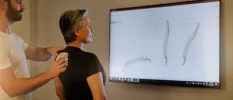 Woman in a black t shirt watching a screen showing images of the spine as a healthcare expert runs a machine down her black