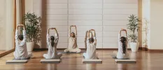 Group of people sit on cushions with their hands stretched up and clasped together as an instructor demonstrates from the front, in a studio with white walls and a wooden floor