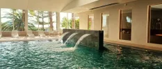 Indoor pool with water jets, surrounding white loungers and full-length window with views of tropical gardens