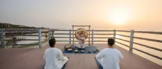 Man and woman in white enjoy a sound healing class on a wooden deck as the sun sets behind the ocean