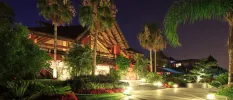 Red building with triangular thatched rooftop surrounded by greenery, lit up under a night sky