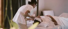 Woman lying down under white bedding while a therapist gives her a facial