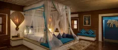 Bedroom with a four-poster bed with a stone base draped in sheer white fabric, with a wooden ceiling, tiled floors and Hindu imagery on the walls