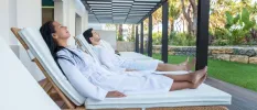 Woman and man in white robes relax on white loungers in a garden area
