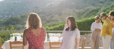 Group of women in summer clothes socialise on a terrace overlooking green hills, as the sun starts to dip beyond them