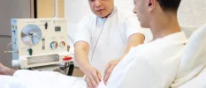 Staff member in white uniform assesses a man lying on a bed, hooked up to a clinical-style machine behind