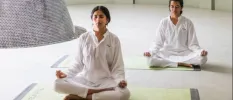 Two women in white sit cross-legged with eyes closed on yoga mats in a white room