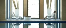 Woman relaxes in a white robe in a chair next to an indoor pool and floor-to-ceiling windows