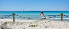 A woman dressed in white dress and hat walking on white sandy beach with crystal clear sea