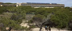 Three people ride horses on a sandy beach backed by a belt of greenery, with a hotel looking down on the scene