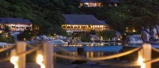 Thatched-roof buildings on the beachfront at night, lit by lanterns and backed by a thick green hillside