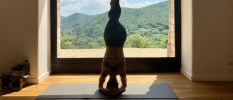 Yoga teacher doing a headstand on a black mat in front of a large window 
