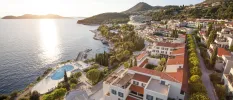 Terracotta-roofed white buildings overlooking the sea, with a swimming pool, grounds dotted with cypress trees and green hills in the background