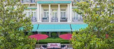Grand facade on a mansion-style building, with a green and white-striped awning, red umbrellas and trees to the front
