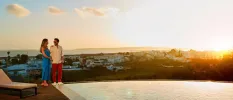 Woman in a blue dress and man in red trousers and a white shirt smile at each other on a glass-fronted rooftop as the sun sets in the background
