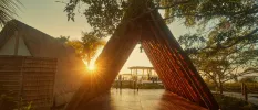 A-frame thatched structure opening onto a wooden deck surrounded by greenery as the sun sets in the background