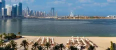 White-sand beach neatly lined with white parasols and loungers, backed by palm trees and with a view of the sea and Dubai skyline