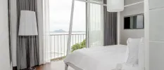 Hotel room with white bedding, grey curtains, white lamp shades and a full-length window with sea views