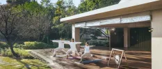 Three people in loose fitting white shirts practice yoga on a wooden deck, overlooking a sunny garden