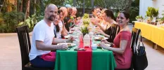 Smiling group pose for a photo while sitting around a communal dining table with a green and red tablecloth and tropical greenery in the background