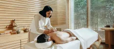 Man lies on his back draped in a white towel as a therapist in white massages him, the treatment room is wood-panelled and has a Buddha statue and sound bowl on a table 