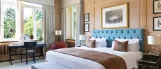 Elegant hotel room with teal headboard, white bedding, a dark-wood dressing table, and pictures of historical images on the wall