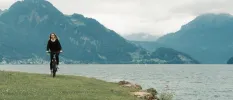 Woman in black cycling along a grassy bank on a lakeside